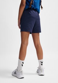 Marina blå sportshorts med elastisk midja, i kombination med vita strumpor med svarta ränder och vita sneakers, sedd från baksidan.