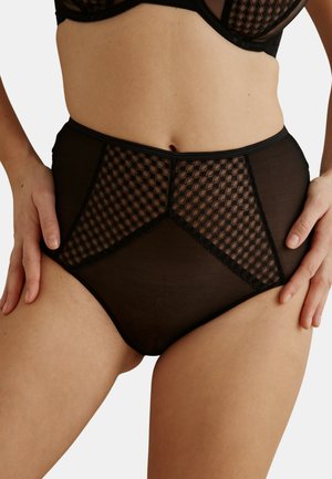 Femme portant une culotte de lingerie noire taille haute avec des panneaux transparents et un motif en mesh géométrique, touchant légèrement ses hanches sur un fond uni.