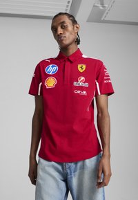 Tricou polo roșu cu guler, având accente albe și mai multe logo-uri, inclusiv Ferrari și Shell. Fabricat dintr-un amestec de bumbac deosebit de moale.
