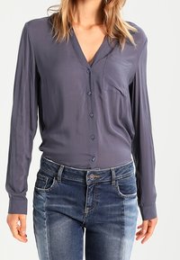 Blouse à manches longues et décolleté en V de couleur gris foncé, dotée d'une poche sur la poitrine et d'une fermeture à bouton. Associée à un jeans en denim bleu délavé.