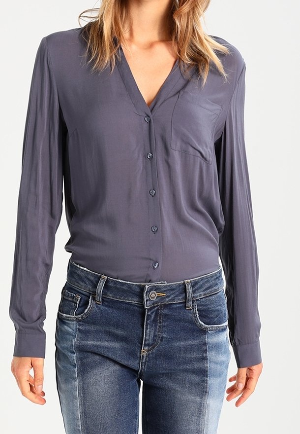 Blouse à manches longues et décolleté en V de couleur gris foncé, dotée d'une poche sur la poitrine et d'une fermeture à bouton. Associée à un jeans en denim bleu délavé.