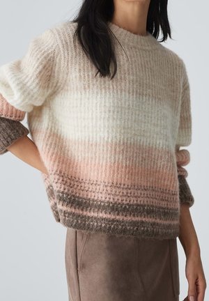 Femme portant un pull en maille duveteuse à rayures horizontales dans des tons crème, rose et marron, associé à un pantalon taupe.