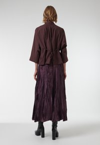 Bruine korte mouw jacket met een strik taille, gecombineerd met een lange, geplooide paarse rok en zwarte platformlaarzen. Gepolijste, gestructureerde materialen.