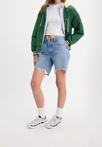 Bomber jacket verde con acentos crema, camiseta corta blanca, pantalones cortos de mezclilla azul claro con dobladillos deshilachados y zapatillas deportivas blancas con acentos negros.