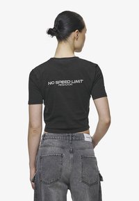 Melnā īsajā krekliņā ar īsām piedurknēm aizmugurē ir balta izšuvuma uzraksts "NAV ĀTRUMA IEROBEŽOJUMU". Apvienots ar augstās jostasvietas pelēkiem džinsiem.