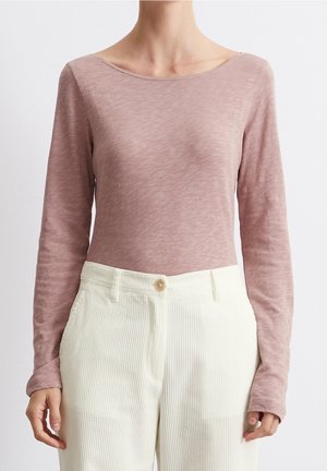 Vrouw draagt een stoffig roze top met lange mouwen, ingestopt in hooggetailleerde, off-white corduroy broek met een enkele knoopsluiting.