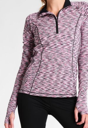 Kvinde iført en langærmet pink og sort mønstret sportstop med tommelhul og sorte leggings, stående mod en ensfarvet baggrund.