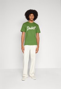 Green cotton T-shirt with white "Doublet" logo, round neckline, and short sleeves, paired with cream wide-leg pants and light sneakers.