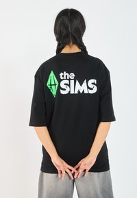Svart oversized T-skjorte med grønt diamantlogo og "the SIMS"-tekst i hvitt på baksiden. Laget av myk, pustende stoff.