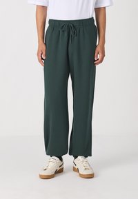 Green sweatpants with an elastic waistband and drawstring detail, featuring a relaxed fit and soft, smooth texture. Worn with white sneakers.