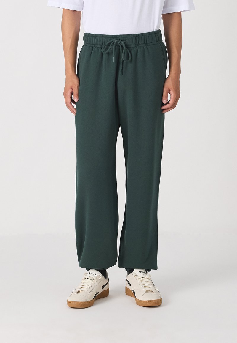 Green sweatpants with an elastic waistband and drawstring detail, featuring a relaxed fit and soft, smooth texture. Worn with white sneakers.