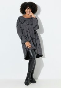 Donna sorridente con capelli ricci e occhiali che indossa un top oversize grigio con stampa di pipistrelli, leggings in pelle neri e stivali neri con borchie.