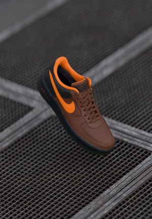 Baskets Nike Air marron et orange avec une semelle noire suspendues au-dessus d'un sol en grille métallique, mettant en valeur le logo latéral et les détails des lacets.