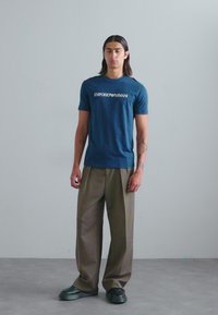 Temno modra bombažna majica s logotipom "EMPORIO ARMANI", v kombinaciji s svetlo rjavimi širokimi hlačami in črnimi masivnimi čevlji. Preprost ozadje.