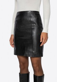 Mini gonna nera in pelle con una texture liscia, silhouette aderente e dettagli sottili di cuciture, abbinata a un top a maniche lunghe di colore chiaro.