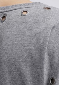 Sudadera gris con ojales metálicos a lo largo de la costura del hombro, textura de tela suave y detalle de cuello acanalado.