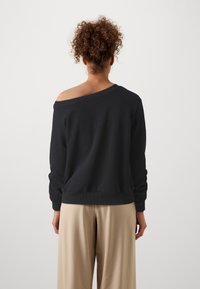 Felpa nera senza spalle con polsini e orlo a coste. Tessuto morbido, vestibilità comoda e design minimalista. Abbinata a pantaloni beige.