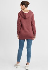 Sweatshirt bordeaux avec une doublure beige douce à l'intérieur de la capuche, doté de manches longues, d'une poche kangourou et d'une finition arrondie, associé à un jean bleu.
