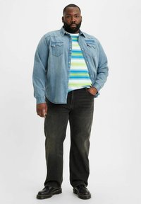 Man som står med ena handen i fickan, iförd en ljusblå denimskjorta över en randig t-shirt i blått, grönt och vitt, svarta jeans och svarta skor.