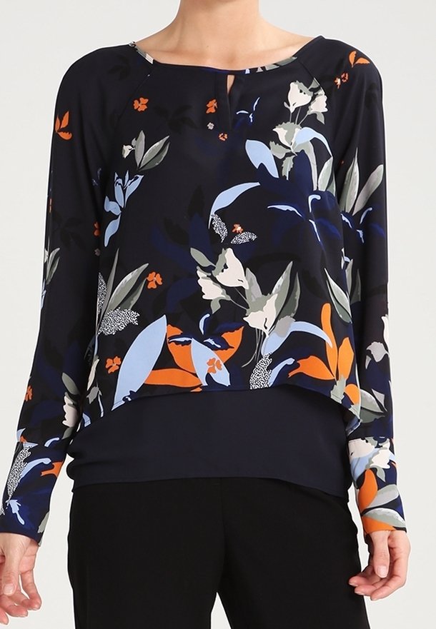 Femme portant un chemisier à manches longues bleu foncé avec de grands motifs floraux orange, bleu clair, beige et gris, associé à un pantalon noir.