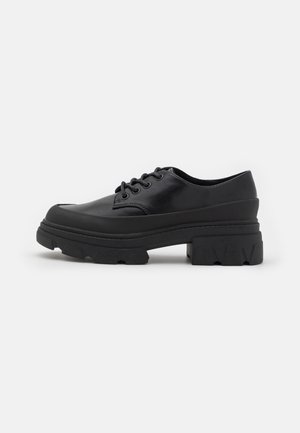Chaussure noire en cuir à lacets avec une semelle épaisse et chunky et un profond profil d'adhérence, conçue pour la durabilité et le style, présentée sur un fond blanc.