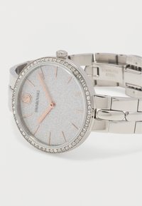 Ασημένιο ρολόι χειρός Swarovski με λαμπερό καντράν, δείκτες και αριθμούς από ροζ χρυσό, στεφάνι διακοσμημένο με κρύσταλλα και κούμπωμα από ανοξείδωτο ατσάλι.