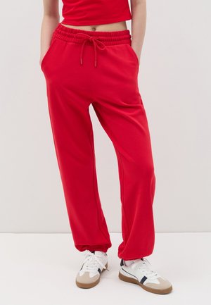 Red sweatpants with an elastic waistband and drawstring, featuring side pockets and cuffed hems, paired with white and beige sneakers.