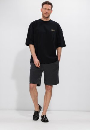 Vīrietis, kurš valkā pārlielu melnu T-kreklu ar uzrakstu "American Vintage", tumši pelēkas šortus un melnas loafer kurpes, stāvot pret baltu fona.