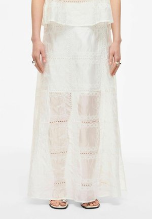 Femme portant une jupe longue en dentelle blanche transparente avec des motifs floraux, debout les mains le long du corps, montrant des bagues et des sandales à bout ouvert.