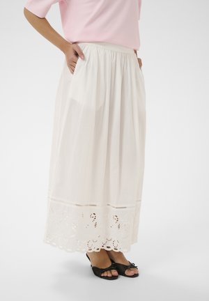 Femme portant une jupe blanche longueur cheville avec un ourlet brodé et des poches, associée à des ballerines noires à bouts ouverts et un haut rose clair.