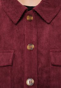 Bordowa kurtka koszulowa z korduroy z klasycznym kołnierzykiem, wyposażona w dwie przednie kieszenie i okrągłe guziki o unikalnym wzorze.