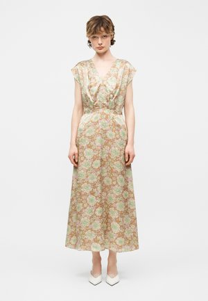 Kvinde iført en blomsterprintet midi-kjole med kappelignende ærmer og v-hals, stående, kombineret med hvide spidse hæle mod en hvid baggrund.