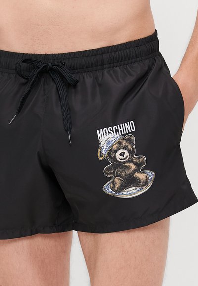 Shorts de bain noirs avec une taille élastique, un cordon de serrage, et un motif d'ours en peluche. "Moschino" est imprimé au-dessus de l'ours.