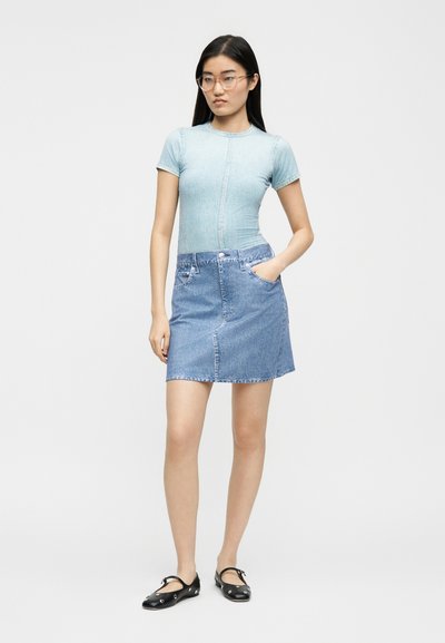 Νεαρή γυναίκα με μακριά σκούρα μαλλιά που φορά γυαλιά, ένα στενό ανοιχτό μπλε μπλουζάκι, φούστα από τζην και μαύρα φλατ παπούτσια με λουράκια στον αστράγαλο.