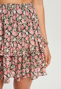 Jupe florale présentant un fond noir avec des roses roses et des feuilles vertes. Le tissu est léger avec un design de bordure en volants superposés.
