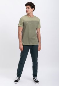 T-shirt verde oliva a maniche corte con stampa grafica vicino al petto, abbinata a pantaloni jogger blu scuro con dettagli in cucitura rossa.