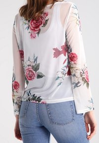 Průsvitná květinová halenka s dlouhými rukávy, bílým základem s růžovými a zelenými květinovými vzory a volným střihem u lemu.