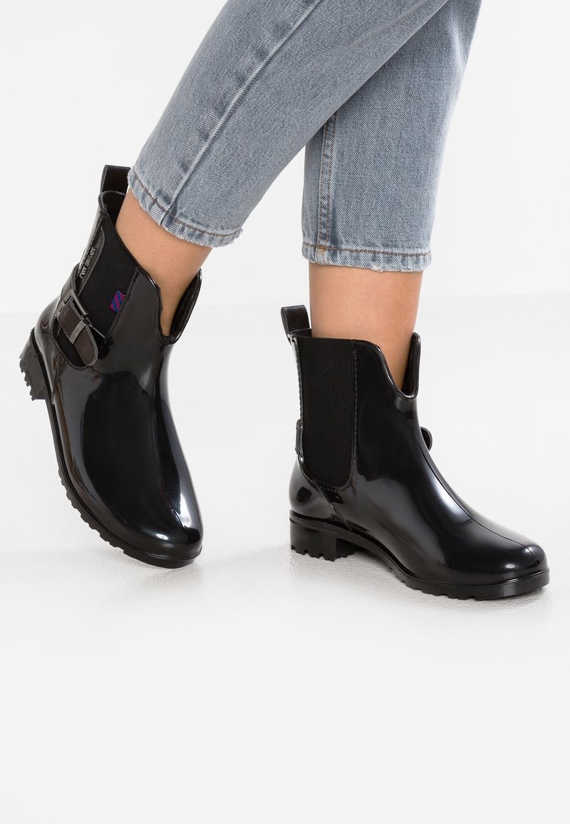Botas de tobillo de goma negras con un acabado brillante, paneles laterales elásticos y un acento de hebilla. Suela plana y una textura en la suela para mayor agarre.