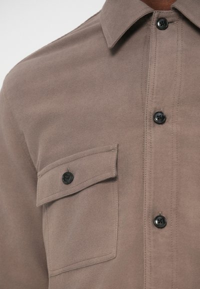 Brun skjorte laget av mykt stoff med knapper foran, svarte knapper og en brystlomme med klaff.