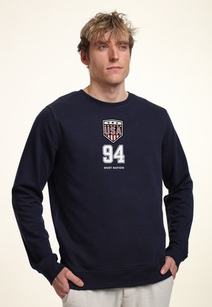 Młody mężczyzna z rozczochranymi blond włosami, ubrany w granatową bluzę z nadrukiem "USA," "94" i "HOST NATION" z przodu.