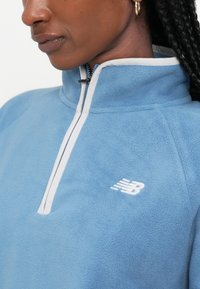 Pulover din polar de culoare albastru deschis, cu guler înalt, fermoar alb și logo brodat alb pe piept. Textură moale și design sportiv.