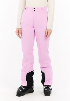 Vrouw in roze skibroek en zwarte skischoenen, staand tegen een witte achtergrond met ontspannen handen langs haar zij.