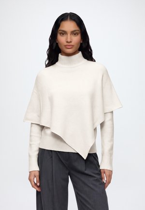 Mujer con cabello largo y negro que lleva un suéter poncho blanco asimétrico y pantalones gris oscuro, de pie sobre un fondo liso.