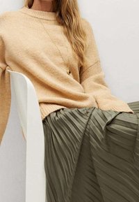 Femme assise sur une chaise blanche portant un pull en maille beige, un collier pendentif en or et une jupe plissée vert olive.