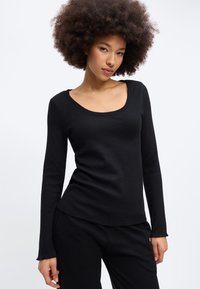 Γυναίκα με σγουρά μαλλιά που φοράει μαύρο μακρυμάνικο τοπ και μαύρο παντελόνι, ποζάροντας μπροστά σε ένα απλό λευκό φόντο.