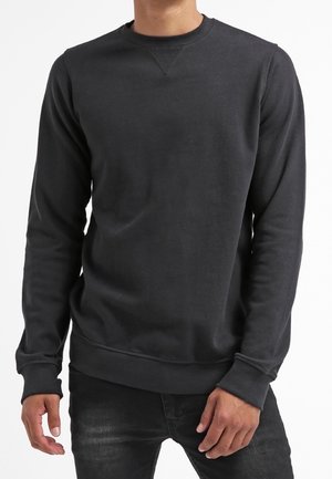 Hombre vistiendo una sudadera de cuello redondo gris oscuro lisa con mangas largas y puños y dobladillo acanalados.