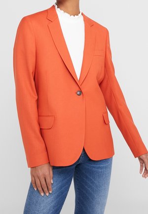 Person trägt ein tailliertes oranges Sakko mit einem schwarzen Knopf, kombiniert mit einem weißen Oberteil und blauen Jeans vor einem einfarbigen Hintergrund.