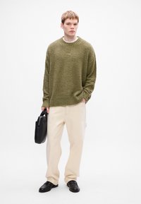 Maglione chunky verde oliva a maglia, pantaloni crema larghi, borsa nera, scarpe slip-on nere; tessuto testurizzato, orlo a coste, vestibilità rilassata.