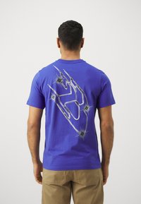 Camiseta de algodón azul con un gráfico metálico plateado de una mano y acentos de estrellas en la parte posterior. Diseño de manga corta y cuello redondo.