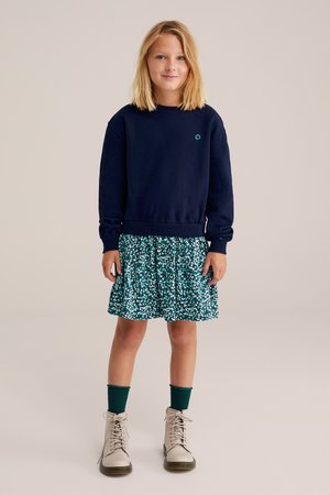 Mörkblå sweatshirt med ribbade muddar, i kombination med en grön blommig klänning med utsvängd kjol. Ljust beige snörkängor och gröna strumpor.
